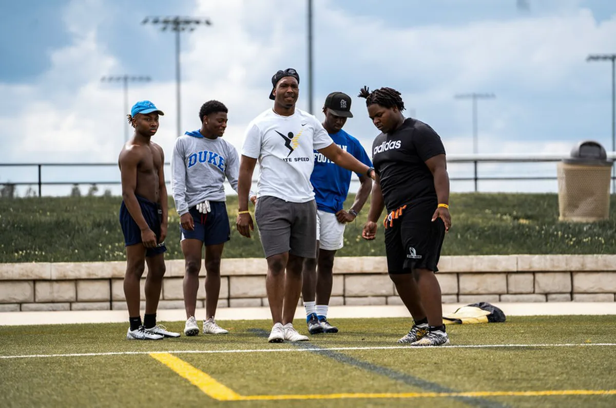An Elite Speed coach in a branded shirt working with a group of young football athletes on the outdoor turf field